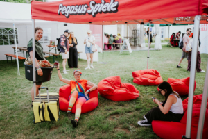 Farbenfrohe Teilnehmer des Elbenwald Festivals entspannen in roten Sitzsäcken unter dem Pegasus Spiele Zelt. Ein Mann mit einem bunten Outfit und einer Kiste steht lächelnd neben einer Frau, die auf ihrem Handy tippt. Festivalkultur und Gemeinschaftsgefühl.