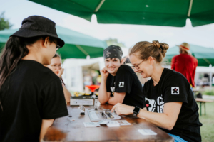 Dice Club Cottbus Gruppe von Teilnehmern beim Elbenwald Festival, die an einem Tisch Brettspiele spielen. Die Atmosphäre ist entspannt und freundlich, mit grünen Zelten im Hintergrund, die den Bereich beschatten. Die Freude am Spiel und am Austausch ist deutlich spürbar.