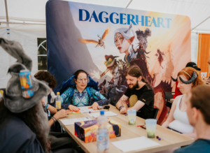 Dice Club Cottbus Eine Gruppe von Teilnehmern am Elbenwald Festival sitzt an einem Tisch und spielt ein Rollenspiel. Im Hintergrund ist ein großes Banner mit dem Titel "Daggerheart" und fantasievollen Charakteren zu sehen. Die Atmosphäre ist kreativ und gemeinschaftlich.