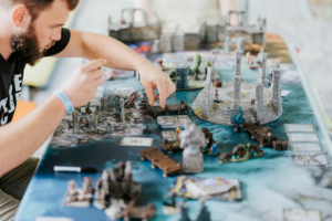 Dice Club Cottbus Ein Teilnehmer des Elbenwald Festivals konzentriert sich auf ein Tabletop-Spiel, während er Miniaturen und Karten strategisch auf einem detaillierten Spielbrett anordnet, um ein spannendes Abenteuer zu erleben.