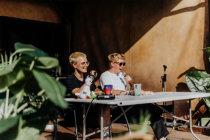 Distrikt 14 Zwei Moderator:innen sitzen an einem Tisch auf dem Elbenwald Festival, umgeben von Pflanzen. Die Person links hat blonde Haare und trägt eine schwarze T-Shirt, während die Person rechts in Sonnenbrille spricht und Notizen macht. Ein Plüschspielzeug und Becher stehen auf dem Tisch.