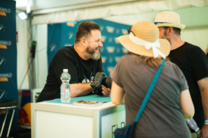 eosAndy Ein lächelnder Mann im T-Shirt steht an einem Tisch und zeigt einer Frau mit Strohhut eine Kamera. Im Hintergrund sind Banner des Elbenwald Festivals zu sehen, die die nerdige Atmosphäre des Events unterstreichen.