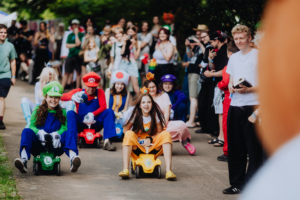 Bunte Cosplayer im Kostüm von Videospielcharakteren, darunter Mario und Luigi, fahren auf kleinen Autos beim Elbenwald Festival. Die fröhliche Atmosphäre zieht eine große Menge von Zuschauern an, die das Spektakel mit Begeisterung verfolgen.