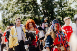 Cosplay auf dem Elbenwald Festival: Eine Frau mit rotem Haar und einer Waffe in der Hand posiert, während eine bunte Gruppe von Festivalbesuchern im Hintergrund vorbeigeht. Der sonnige Tag und die lebhafte Atmosphäre unterstreichen die Vielfalt der Nerd-Kultur.