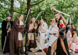 Gruppenszene auf dem Elbenwald Festival mit Teilnehmern in kreativen Kostümen, darunter Jedi-Ritter und verschiedene Charaktere, die mit Lichtschwertern posieren. Im Hintergrund sind weitere Festivalbesucher und grüne Bäume zu sehen, die die lebendige Atmosphäre unterstreichen.