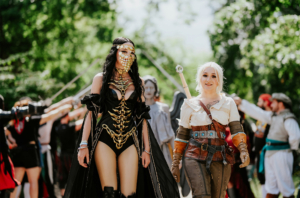 Zwei Frauen in aufwendigen fantastischen Kostümen gehen über das Elbenwald Festival. Die erste trägt ein schauriges Outfit mit goldenen Akzenten, während die zweite in einem abenteuerlichen Look mit Schwert und Gürtel gekleidet ist. Im Hintergrund sind weitere Cosplayer zu sehen.