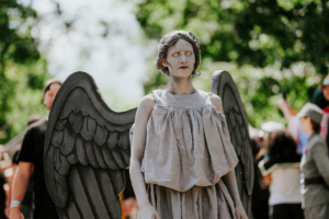 Eine verkleidete Person mit Engelsflügeln und blasser Haut steht im Vordergrund des Elbenwald Festivals, umgeben von einer lebhaften Festivalatmosphäre. Die Kostümierung erinnert an eine mystische Figur und zieht die Aufmerksamkeit der Besucher auf sich.