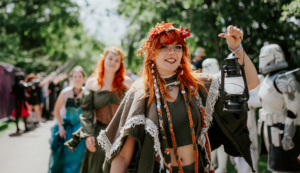 Eine fröhliche Teilnehmerin des Elbenwald Festivals trägt eine Laterne und zeigt ihren kreativen Kostümstil mit roten Haaren und Blumenkranz. Im Hintergrund sind weitere verkleidete Festbesucher und Kostüme zu sehen, die die festliche Atmosphäre unterstreichen.