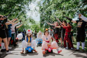 Zwei Cosplayer in bunten Spielzeugautos, gekleidet als Toad und Prinzessin Peach, sitzen auf einem Festivalgelände umgeben von Menschen in Kostümen, die Schwerter in die Höhe halten. Die Szene strahlt eine fröhliche, nerdige Atmosphäre aus.