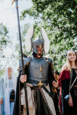 Ein beeindruckender Cosplayer in aufwendiger Rüstung mit Flügelhelm und Stab, umgeben von anderen Teilnehmern des Elbenwald Festivals. Die Atmosphäre ist lebhaft, mit grünen Bäumen im Hintergrund, die das Festival-Feeling unterstreichen.