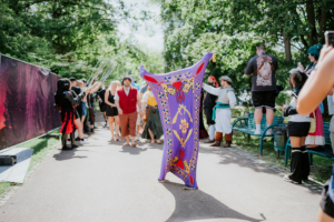 Farbenfrohe Cosplay-Szene beim Elbenwald Festival, mit Teilnehmern in fantasievollen Kostümen, die auf einem Weg stehen. Ein Teilnehmer hebt ein kunstvolles Banner, während andere in historischen und fantastischen Outfits posieren. Grünflächen und festliche Atmosphäre im Hintergrund.