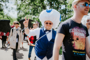 Ein Teilnehmer des Elbenwald Festivals trägt ein auffälliges Kostüm mit einem großen, weißen Hut und einer blauen Verzierung. Im Hintergrund sind weitere kostümierte Festivalbesucher zu sehen, die die lebendige Atmosphäre des Events widerspiegeln.