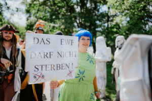 Frau mit blauer Perücke und grünem Kleid hält ein Plakat mit der Aufschrift "Das EWF darf nicht sterben!" beim Elbenwald Festival. Im Hintergrund sind verkleidete Teilnehmer, darunter ein Pirat und Stormtrooper, zu sehen.