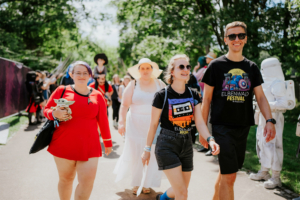Vier Festivalbesucher beim Elbenwald Festival, gekleidet in kreative Kostüme, gehen fröhlich einen schattigen Weg entlang. Im Hintergrund sind weitere Teilnehmer in bunten Outfits zu sehen, die die lebhafte Atmosphäre des Events unterstreichen.