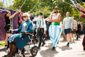 Eine bunte Parade beim Elbenwald Festival: Eine Frau im Rollstuhl mit einem blauen Kleid und einer Blume, umgeben von kostümierten Teilnehmern, darunter ein Mann in einem auffälligen Outfit und andere Festivalbesucher, die die kreative Atmosphäre genießen.