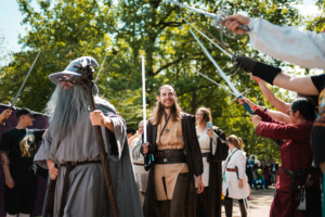Teilnehmer des Elbenwald Festivals in fantasievollen Kostümen, darunter ein Zauberer und Jedi, posieren mit Schwertern vor einer jubelnden Menge. Die Veranstaltung vereint Musik und Nerd-Kultur in einer lebhaften Atmosphäre unter freiem Himmel.