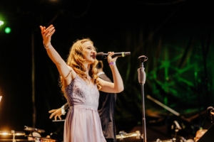 Elbenwald in Concert Eine Sängerin mit langen, lockigen Haaren trägt ein elegantes Kleid und performt leidenschaftlich auf der Bühne beim Elbenwald Festival. Im Hintergrund sind Musiker mit Instrumenten zu sehen, während die Lichter der Veranstaltung funkeln.
