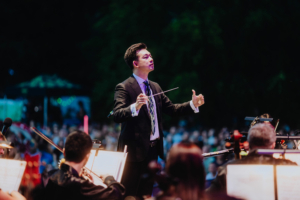 Elbenwald in Concert Dirigent eines Orchesters auf der Bühne beim Elbenwald Festival, umgeben von Musikern, die für ein begeistertes Publikum spielen. Die Atmosphäre vereint musikalische Darbietungen mit der Energie eines Festivals und der Vielfalt von Nerd-Kultur.