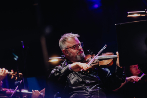 Elbenwald in Concert Ein Musiker spielt Geige auf der Bühne des Elbenwald Festivals, umgeben von anderen Orchestermitgliedern. Die Atmosphäre ist lebhaft und kreativ, während die Lichter die Bühne erhellen und die Musik das Publikum begeistert.