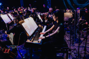 Elbenwald in Concert Eine Musikerin spielt auf einem E-Piano während des Elbenwald Festivals, umgeben von einem Orchester. Die Bühne ist mit bunten Lichtern beleuchtet, während Notenblätter und Instrumente im Vordergrund zu sehen sind, was die kreative Atmosphäre des Events unterstreicht.