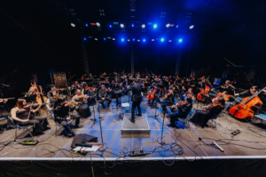 Elbenwald in Concert Ein Orchester spielt auf der Bühne des Elbenwald Festivals, umgeben von bunten Lichtern. Die Musiker in eleganten Outfits konzentrieren sich auf ihre Instrumente, während der Dirigent das Ensemble leitet. Eine einzigartige Verbindung von Musik und Nerd-Kultur.