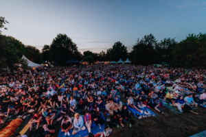 Elbenwald in Concert Ein großer Menschenauflauf beim Elbenwald Festival, das eine Mischung aus Musikfestival und Nerd-Convention ist. Die Zuschauer sitzen auf Decken im Freien, umgeben von Bäumen und Zelten, während der Abendhimmel den Veranstaltungsort beleuchtet.