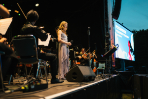 Elbenwald in Concert Eine Sängerin steht auf der Bühne des Elbenwald Festivals und performt einen Song, während ein Orchester sie begleitet. Im Hintergrund ist ein Bildschirm zu sehen, der den Titel "Pocahontas - Colours of the Wind" anzeigt.