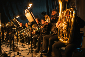 Elbenwald in Concert Musiker auf der Bühne des Elbenwald Festivals, die verschiedene Blasinstrumente spielen. Die Band ist in eleganter Kleidung gekleidet und sorgt für eine festliche Stimmung mit einer Mischung aus Musik und nerdiger Kultur.