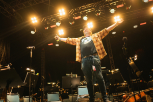 Ein begeisterter Moderator steht auf der Bühne des Elbenwald Festivals, umgeben von Mikrofonen und Musiknoten. Die Scheinwerfer strahlen hell, während er mit offenen Armen das Publikum anfeuert und die energiegeladene Atmosphäre des Events verkörpert.
