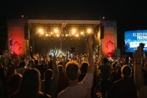 Menschenmenge beim Elbenwald Festival, mit erhobenen Händen und bunten Lichtern auf der Bühne. Die Atmosphäre ist lebhaft und feierlich, während die Festivalbesucher das Musik- und Konventionserlebnis genießen. Datum: 6-8. August.