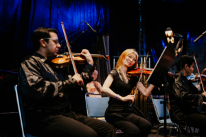 Elbenwald in Concert Musiker spielen Geige auf der Bühne des Elbenwald Festivals, umgeben von einer bunten Beleuchtung. Die Bandmitglieder tragen elegante schwarze Outfits und konzentrieren sich auf ihre Performance, während das Publikum gespannt zuschaut.