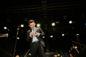 Elbenwald in Concert Ein lächelnder Moderator in Anzug hält ein Mikrofon und Notizen auf der Bühne des Elbenwald Festivals, umgeben von Scheinwerfern und einem Orchester. Die Atmosphäre ist lebhaft und energiegeladen, während das Publikum gespannt zuhört.