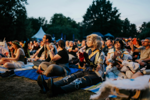 Elbenwald in Concert Besucher des Elbenwald Festivals sitzen auf einer Wiese und genießen die Atmosphäre. Einige sind in aufwendigen Kostümen gekleidet, während andere entspannt applaudieren. Die Mischung aus Musik und Gemeinschaft schafft eine lebendige, festliche Stimmung.