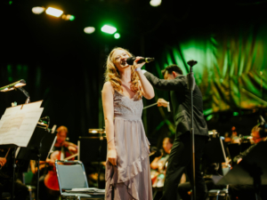 Elbenwald in Concert Sängerin auf der Bühne des Elbenwald Festivals, umgeben von Orchestermusikern. Die Atmosphäre ist lebhaft, mit bunten Lichtern und einem engagierten Publikum, das die Mischung aus Musik und Nerd-Kultur genießt.