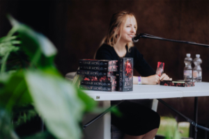Elena Zay Autorin bei der Buchsignierung auf dem Elbenwald Festival, umgeben von ihren Werken "Night Jewel". Im Hintergrund sind Wasserflaschen und eine grüne Pflanze sichtbar, während die Autorin am Mikrofon spricht.