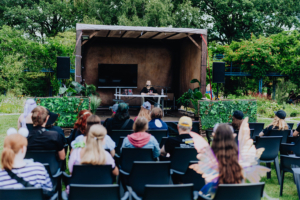 Elena Zay Auf der Bühne des Elbenwald Festivals spricht eine Moderatorin vor einem Publikum in Kostümen. Im Hintergrund ist ein Bildschirm und eine grüne Pflanzenwand zu sehen. Die Veranstaltung vereint Musik und Nerd-Kultur in einer einzigartigen Atmosphäre.