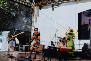 Endgegner Musiker auf der Bühne des Elbenwald Festivals, gekleidet in fantasievolle Kostüme. Die Band spielt live, umgeben von Instrumenten und einer festlichen Atmosphäre. Im Hintergrund ist das Festival-Logo sichtbar, das die nerdige Kultur feiert.