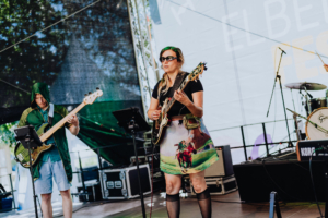 Endgegner Bandauftritt auf dem Elbenwald Festival: Musikerin mit Gitarre und bunter Rock, Bassist im grünen Outfit auf der Bühne. Die Atmosphäre verbindet Musik und Nerd-Kultur, umgeben von Festival-Feeling und kreativen Kostümen.