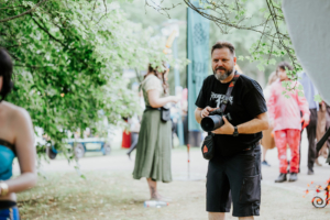 eosAndy Ein Fotograf mit Kamera steht im Vordergrund des Elbenwald Festivals, umgeben von Besuchern in fantasievoller Kleidung und einer grünen, lebendigen Umgebung. Die Atmosphäre ist lebhaft und kreativ, perfekt für Fans von Musik und Nerd-Kultur.