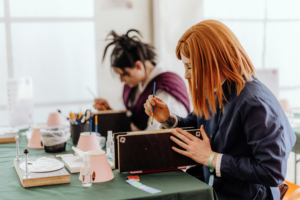 Farbschnitt Zwei Teilnehmer des Elbenwald Festivals sitzen an einem Tisch und malen an ihren Kunstprojekten. Auf dem Tisch sind Malutensilien, Wasserbehälter und kreative Materialien sichtbar, während die Atmosphäre kreativ und entspannt wirkt.