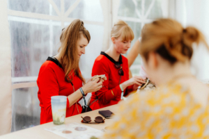 Feenhandwerk Zwei Frauen in roten Kostümen arbeiten an Bastelprojekten während des Elbenwald Festivals. Auf dem Tisch liegen Sonnenbrillen und ein Getränk. Die Atmosphäre ist kreativ und freundlich, ideal für Fans von Musik und Nerd-Kultur.