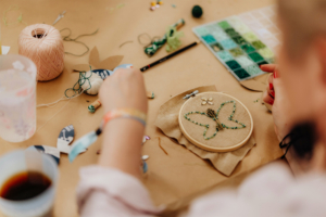 Feenhandwerk Eine Person arbeitet an einer handgefertigten Stickerei eines Schmetterlings mit grünen Perlen und goldenen Akzenten. Um sie herum liegen Nähmaterialien, Farben und Bastelutensilien, die die kreative Atmosphäre des Elbenwald Festivals widerspiegeln.