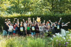 Fellowship Area Eine Gruppe von Festivalbesuchern beim Elbenwald Festival, die fröhlich in einer blühenden Umgebung posieren. Sie tragen kreative Kostüme und T-Shirts, während einige eine Pikachu-Plüschfigur halten. Die Szene strahlt Freude und Gemeinschaftsgeist aus.