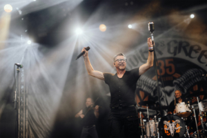 Fiddler's Green Ein Sänger mit Brille und Mikrofon auf der Bühne des Elbenwald Festivals, umgeben von Lichtern und Nebel. Im Hintergrund sind Musiker mit Instrumenten zu sehen, die die energiegeladene Atmosphäre des Festivals widerspiegeln.