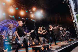 Fiddler's Green Live-Auftritt der Band auf dem Elbenwald Festival, mit energiegeladenen Musikern auf der Bühne, darunter ein Bassist, ein Geiger und ein Sänger. Die Atmosphäre ist festlich, mit bunten Lichtern und einer beeindruckenden Bühnenkulisse.