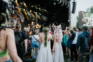 Zuschauer genießen Live-Musik auf dem Elbenwald Festival, umgeben von bunten Kostümen und Seifenblasen. Die Atmosphäre ist lebhaft, während Künstler auf der Bühne auftreten und das Publikum begeistert mitfeiert.