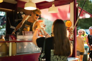 Ein Verkäufer reicht einem Festivalbesucher ein Eis an einem bunten Stand auf dem Elbenwald Festival. Im Hintergrund sind fröhliche Menschen zu sehen, die die Atmosphäre des einzigartigen Events genießen. Leckere Snacks und gute Stimmung prägen die Szene.