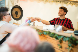 Ein Besucher des Elbenwald Festivals erhält eine Pizza von einem Verkäufer an einem Foodstand. Im Hintergrund ist eine große Glocke sichtbar, die zur Atmosphäre des Festivals beiträgt. Die Szene vermittelt die gesellige Stimmung des Events.