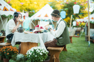 Zwei Festivalbesucher genießen ein Picknick auf dem Elbenwald Festival. Sie sitzen an einem Tisch mit einer weißen Tischdecke, umgeben von bunten Blumen und festlicher Dekoration, während sie Snacks und Getränke teilen.