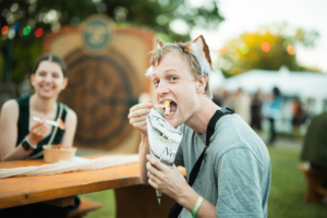 Junger Mann mit Katzenohren genießt Snacks auf dem Elbenwald Festival, während eine lachende Frau im Hintergrund sitzt. Die entspannte Atmosphäre und das bunte Festivalambiente sind deutlich zu erkennen.